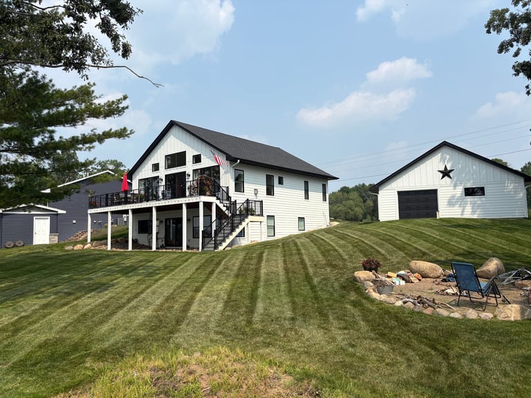 Two-story farmhouse with striped lawn, covered deck, detached garage with star, and landscaped garden on sunny day