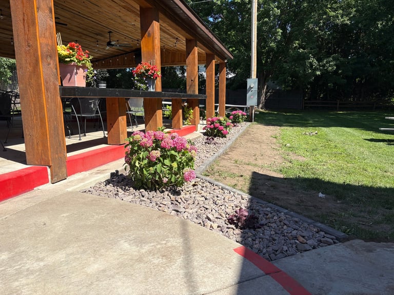 Wooden pavilion with red trim and pink flowers in landscaped beds along a concrete pathway with green lawn in background