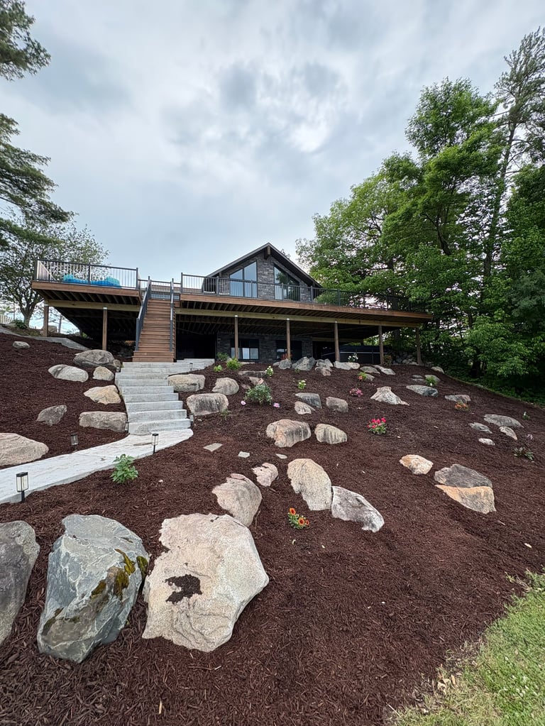 Modern wooden house with stone landscaping features, concrete pathway, and lush green trees surrounding the property on a cloudy day