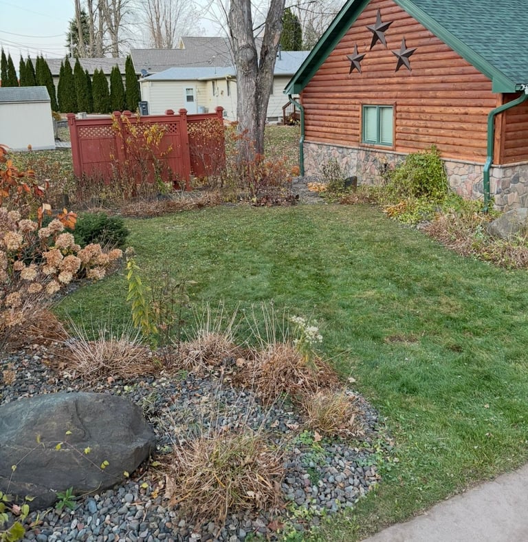 Residential backyard with red barn, grass, dormant brown shrubs, gravel landscaping, and stone foundation in autumn
