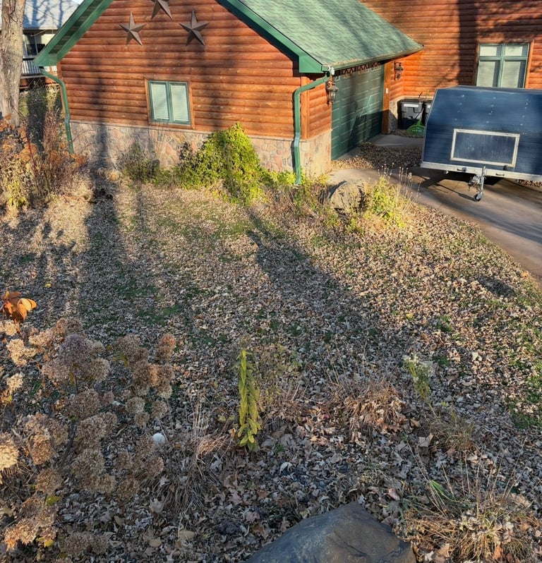 Wooden cabin with green roof on gravel ground, blue trailer visible in background
